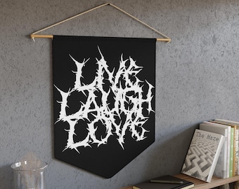 May include: Black banner with the words "LIVE LAUGH LOVE" in white, spiky, gothic-style font. The banner is suspended from a wooden dowel with natural twine. The banner is displayed on a grey wall.