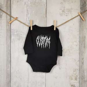 Könnte beinhalten: Schwarzer Baby-Body mit langen Ärmeln und dem weißen Wort "CUSTOM" in einer stilisierten Schriftart. Der Body hängt an einer Wäscheleine mit Holzklammern vor einem hellen Holzgrund.