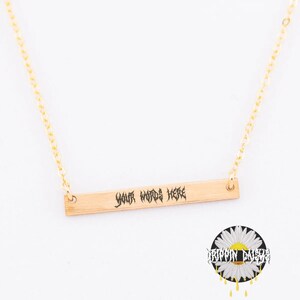 Könnte beinhalten: Eine goldfarbene Halskette mit einem rechteckigen Anhänger, auf dem in gotischer Schrift "YOUR WORDS HERE" steht. Die Kette hängt an einer feinen Goldkette. Ein Gänseblümchen-Logo mit dem Text "DRIPPIN DAISY" ist unten rechts zu sehen.
