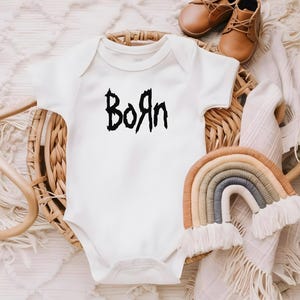 Könnte beinhalten: Ein weißer Baby-Strampler mit schwarzem Text, der "Born" in einer gotischen Schriftart sagt. Der Strampler befindet sich in einem Weidenkorb mit einem regenbogenfarbenen gewebten Wandbehang.
