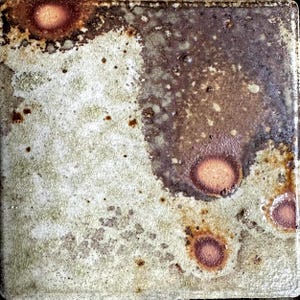 May include: Square ceramic tile with a mottled, textured surface. The tile features a blend of colors, including off-white, brown, and reddish-brown, with a crackle glaze effect. Circular reddish-brown spots are scattered across the surface, adding to the rustic aesthetic.