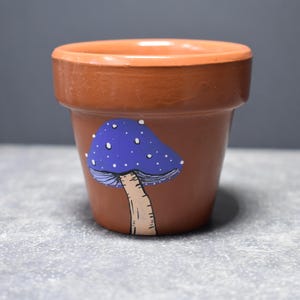 May include: A small, terracotta-colored flower pot with a hand-painted mushroom design. The mushroom has a blue cap with white spots and a beige stem. The pot is unglazed and has a simple, classic shape.