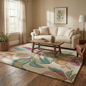 May include: A beige area rug with a large floral design in shades of green, pink, and yellow. The rug is placed in a living room with a cream-colored sofa, wooden coffee table, and a framed botanical print on the wall.
