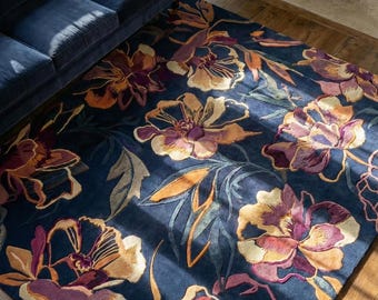 May include: A rectangular area rug with a dark blue background and a large floral pattern in shades of purple, orange, and gold. The rug is placed on a hardwood floor, with a blue sofa visible in the background.