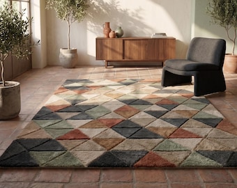 May include: A large area rug with a geometric pattern of triangles in various colors, including beige, green, orange, and gray. The rug is placed on a brick floor, with a chair and wooden cabinet in the background. Olive trees in pots are also visible.
