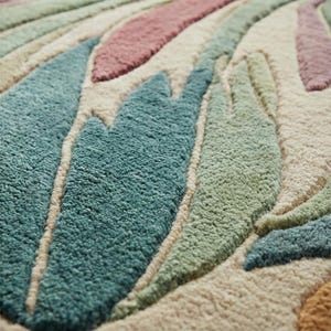 May include: Close-up of a textured rug with a leaf design. The rug features a beige background with large, stylized leaves in shades of teal, green, pink, and peach. The leaves have a raised, textured appearance, creating a soft, inviting look.