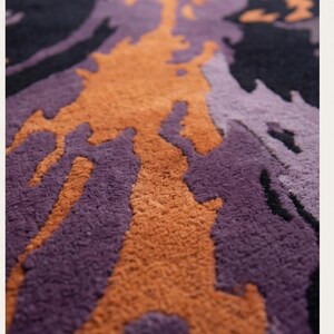 May include: A tufted rug featuring an abstract, organic pattern with deep black, vibrant orange, and various shades of purple. The surface has a thick, textured pile with distinct color-blocked shapes that flow across the entire area of the rug.