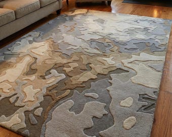 May include: A large area rug with a unique, abstract design in shades of gray, beige, and brown. The rug has a textured, raised pattern that adds depth and visual interest. It is placed on a hardwood floor in a living room setting.