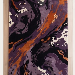 May include: A rectangular area rug featuring an abstract, swirling pattern in shades of deep purple, light lavender, burnt orange, black, and cream. The design consists of fluid, organic shapes that flow across the surface in a dynamic composition.