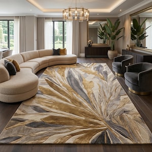 May include: A large rectangular area rug featuring a prominent abstract botanical pattern in shades of gold, beige, and charcoal gray. The design consists of elongated, layered leaf-like shapes that radiate from one corner across the surface.