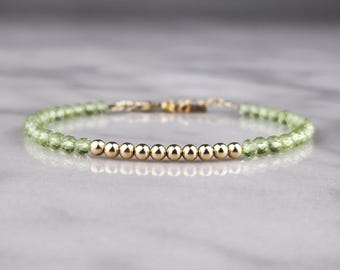May include: Delicate bracelet featuring a strand of small, light green beads interspersed with a row of gold-colored beads. The bracelet is strung on a thin, gold-colored chain, and the clasp is also gold-colored. The bracelet is displayed on a light gray surface.