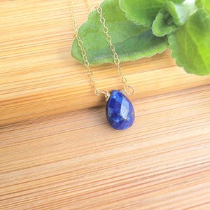 May include: A gold chain necklace featuring a faceted, teardrop-shaped, deep blue gemstone pendant. The necklace is displayed on a wooden surface with a green leaf in the background. The pendant is approximately 2 cm in length.