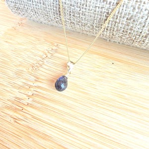 May include: A gold-colored necklace with a teardrop-shaped, dark blue gemstone pendant. The pendant is faceted, catching the light. The necklace is displayed on a wooden surface with a neutral-colored fabric in the background. Jewelry.