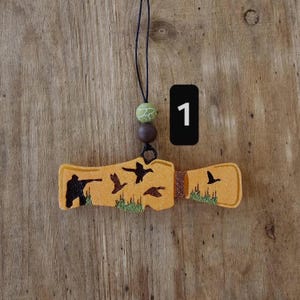 May include: An orange-yellow air freshener with a hunting theme. The design features a hunter, ducks, and grass. The air freshener is suspended by a black cord with green and brown beads. A black rectangle with the number 1 is also present.