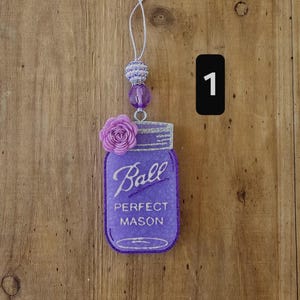 May include: A purple mason jar-shaped ornament with the word "Ball" in script, and "Perfect Mason" below. It has a silver glitter rim and a purple rose accent. The ornament is suspended by a string with decorative beads.