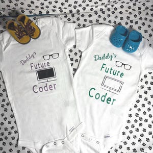 Könnte beinhalten: Zwei weiße Baby-Bodys mit dem Text "Daddy's Future Coder" und einer Computergrafik. Ein Body hat lila Text, der andere grün. Gelbe und blaue Babyschuhe sind in der Nähe der Bodys platziert.