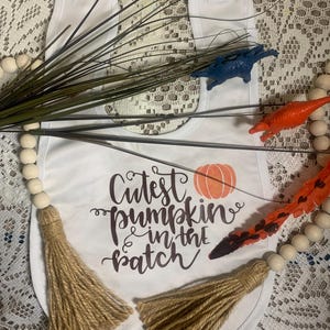 Może przedstawiać: Biały śliniak dla niemowlęcia z napisem "Cutest pumpkin in the patch" i małym pomarańczowym obrazkiem dyni. Śliniak ma dwa brązowe frędzle.