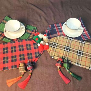 May include: Four placemats with different plaid patterns, two white teacups and saucers, and two small decorative ornaments. The placemats are green, blue, red, and tan.