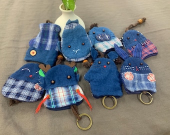 May include: A collection of eight handmade keychains featuring various animal designs. The keychains are made from denim and plaid fabric and feature embroidered details. Each keychain has a metal ring for attaching keys.