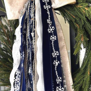 May include: A festive Christmas wreath with a large, cream-colored velvet bow. The wreath is adorned with various ribbons in navy blue, white, and gold, some featuring snowflake and tree patterns. The evergreen branches provide a natural backdrop.