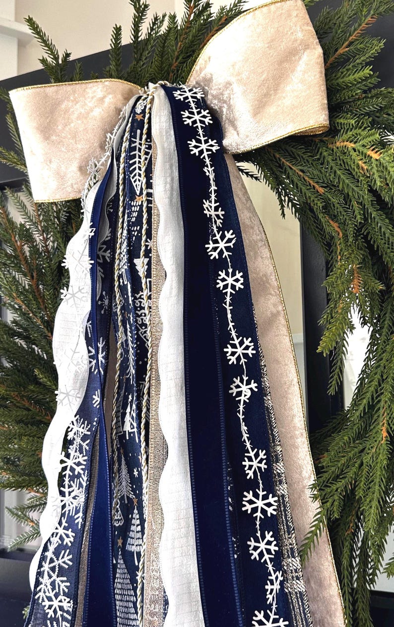 May include: A festive Christmas wreath with a large, cream-colored velvet bow. The wreath is adorned with various ribbons in navy blue, white, and gold, some featuring snowflake and tree patterns. The evergreen branches provide a natural backdrop.