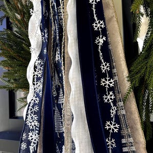 May include: Close-up of decorative ribbons in shades of navy blue, white, and gold. The ribbons feature snowflake and tree patterns, along with a scalloped white ribbon. The ribbons are part of a holiday decoration.