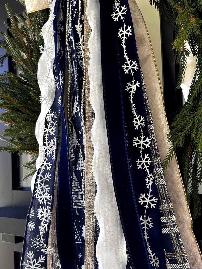 May include: Close-up of decorative ribbons in shades of navy blue, white, and gold. The ribbons feature snowflake and tree patterns, along with a scalloped white ribbon. The ribbons are part of a holiday decoration.