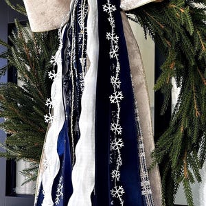 May include: A festive Christmas wreath with a large cream-colored bow with gold trim. The wreath is adorned with evergreen branches and cascading ribbons in navy blue, white, and silver, some decorated with snowflake patterns. A decorative holiday accent.