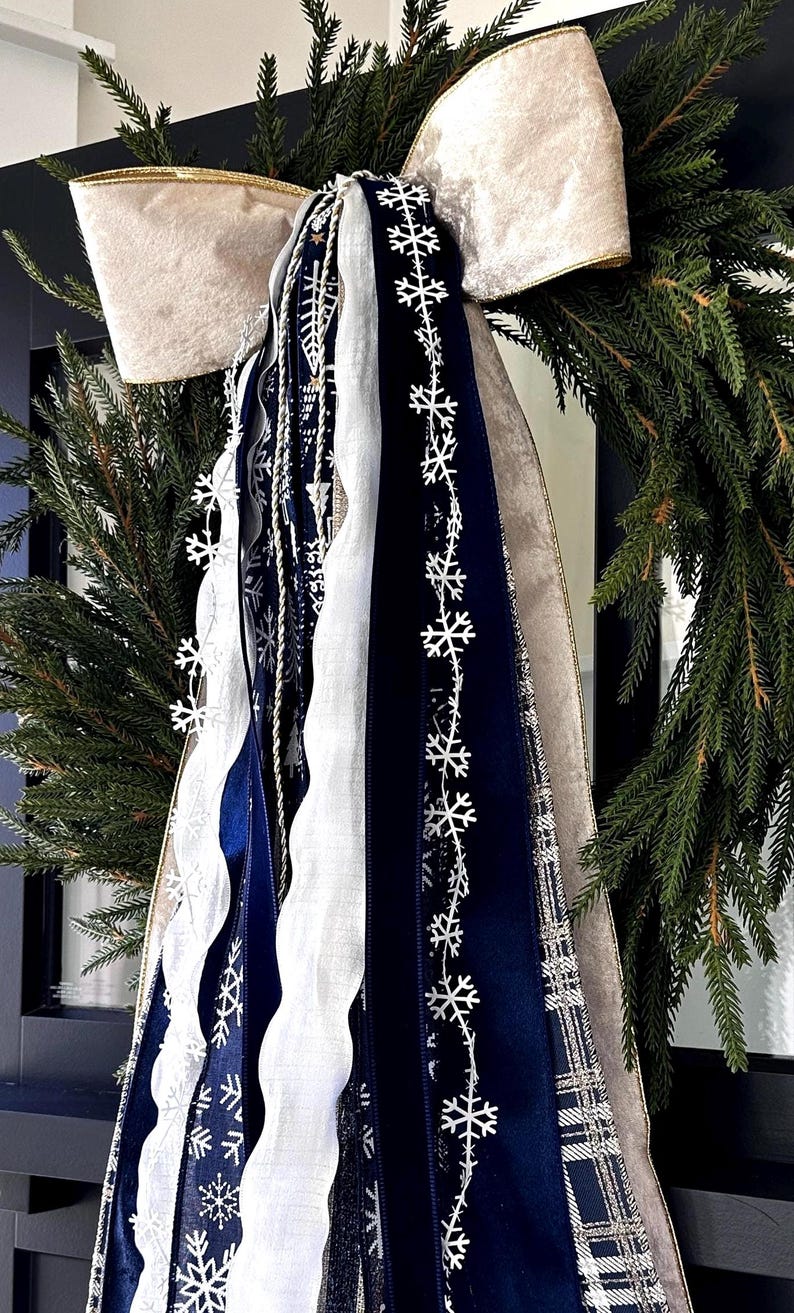 May include: A festive Christmas wreath with a large cream-colored bow with gold trim. The wreath is adorned with evergreen branches and cascading ribbons in navy blue, white, and silver, some decorated with snowflake patterns. A decorative holiday accent.