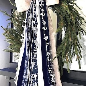 May include: A festive Christmas wreath with lush green pine branches. It features a large cream-colored bow and decorative ribbons in navy blue, white, and cream, adorned with snowflake and holiday-themed patterns. A perfect holiday decoration.