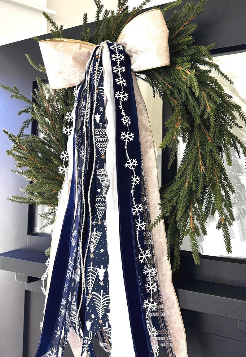 May include: A festive Christmas wreath with lush green pine branches. It features a large cream-colored bow and decorative ribbons in navy blue, white, and cream, adorned with snowflake and holiday-themed patterns. A perfect holiday decoration.