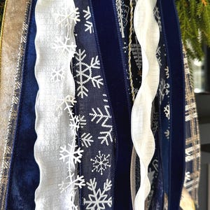 May include: A close-up of various decorative ribbons in shades of blue, white, and gold. The ribbons include a navy blue velvet ribbon with white snowflake patterns, a white ribbon with a wavy edge, and a gold ribbon with a plaid design. The ribbons are likely for holiday decorations.
