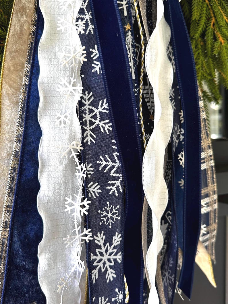 May include: A close-up of various decorative ribbons in shades of blue, white, and gold. The ribbons include a navy blue velvet ribbon with white snowflake patterns, a white ribbon with a wavy edge, and a gold ribbon with a plaid design. The ribbons are likely for holiday decorations.