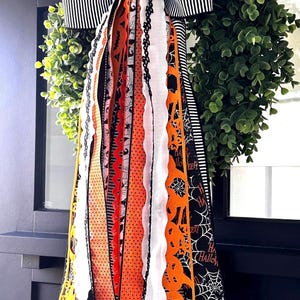 May include: A Halloween-themed wreath with a green foliage base, adorned with a black and white striped bow. Colorful ribbons in orange, black, and white cascade down, featuring spiderwebs, pumpkins, and the text "Happy Halloween."