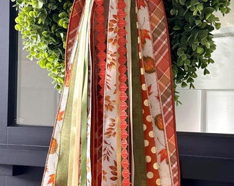 May include: A decorative wreath featuring a dense arrangement of green leaves topped with a large bow made of plaid fabric in shades of orange and brown. Multiple long ribbons hang down, displaying patterns of autumn leaves, polka dots, and solid colors.