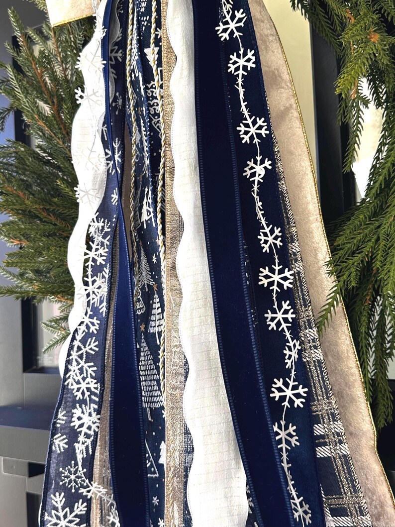 May include: Close-up of decorative ribbons in shades of navy, white, and gold. The ribbons feature snowflake and tree patterns, along with a textured white ribbon and a gold ribbon. The ribbons are part of a holiday decoration.