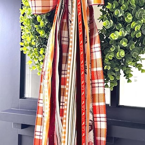 May include: A fall-themed wreath with a green eucalyptus base. A large plaid bow in orange and red accents the top. Various ribbons in coordinating colors and patterns, including one with "Hello Fall" text, cascade down.