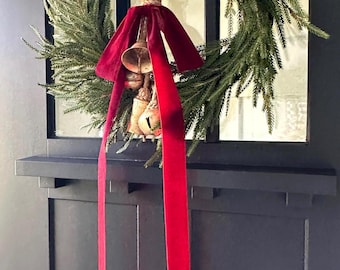May include: A green pine wreath hangs on a dark door, featuring a cluster of metallic bells suspended by a thick jute rope. A long, deep red velvet ribbon is tied in a bow around the rope, with two long tails hanging down over the center.