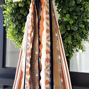 May include: A decorative wreath attachment featuring a large bow with orange and white stripes, cascading down into multiple long fabric ribbons. The ribbons display patterns of autumn leaves, pumpkins, solid brown velvet, and navy blue textures.