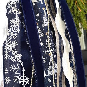 May include: Close-up of decorative ribbons in a winter theme. The ribbons feature snowflake and Christmas tree patterns in white on a navy blue background. Other ribbons include a navy velvet, gold, and white. The ribbons are likely for holiday decor.