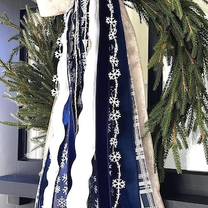 May include: A Christmas wreath with evergreen branches, a large cream-colored bow, and cascading ribbons. The ribbons are navy blue, white, and plaid, with snowflake patterns. The wreath is a festive holiday decoration.