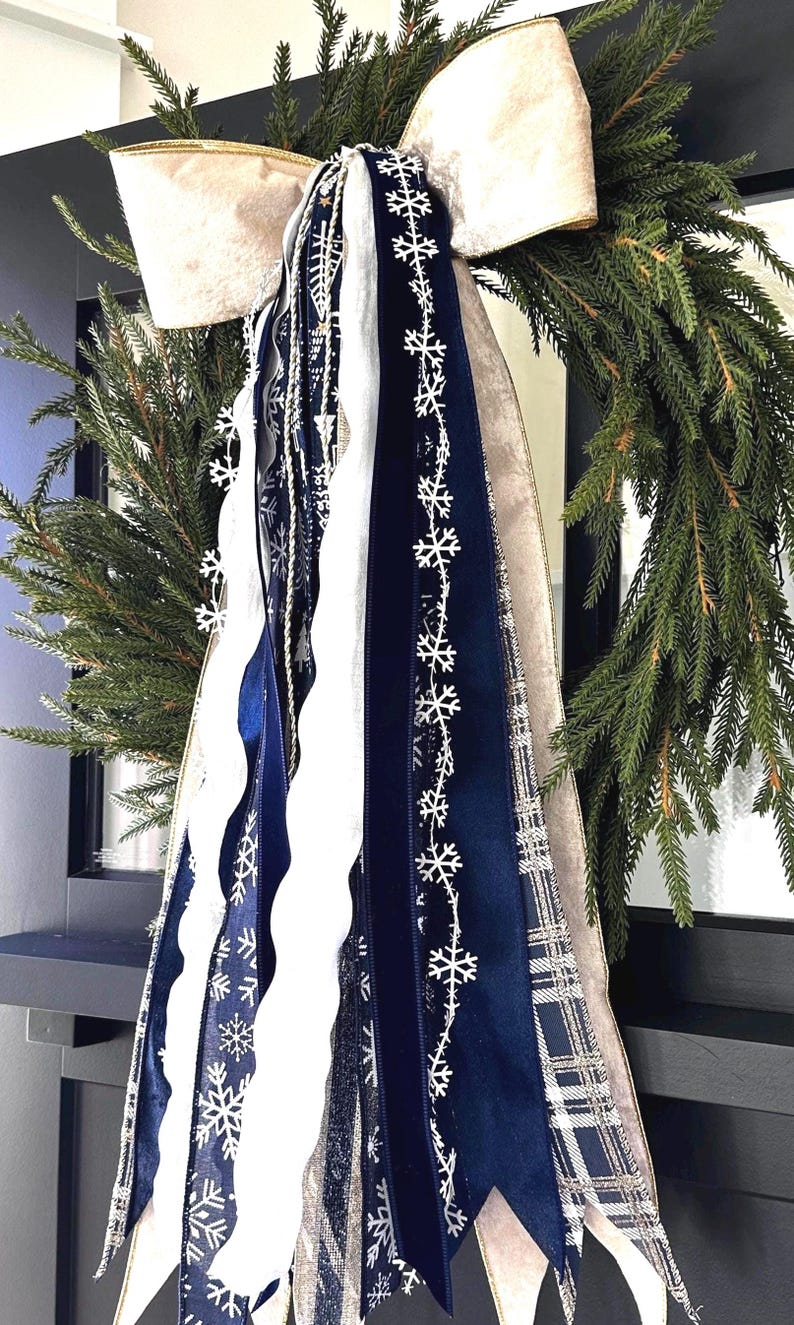 May include: A Christmas wreath with evergreen branches, a large cream-colored bow, and cascading ribbons. The ribbons are navy blue, white, and plaid, with snowflake patterns. The wreath is a festive holiday decoration.