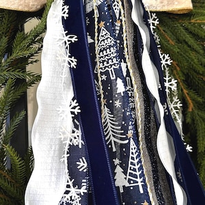 May include: A close-up of a Christmas wreath with various ribbons. The ribbons include white, navy blue, and silver, with snowflake and Christmas tree patterns. The wreath has a gold trim and is surrounded by green pine needles.