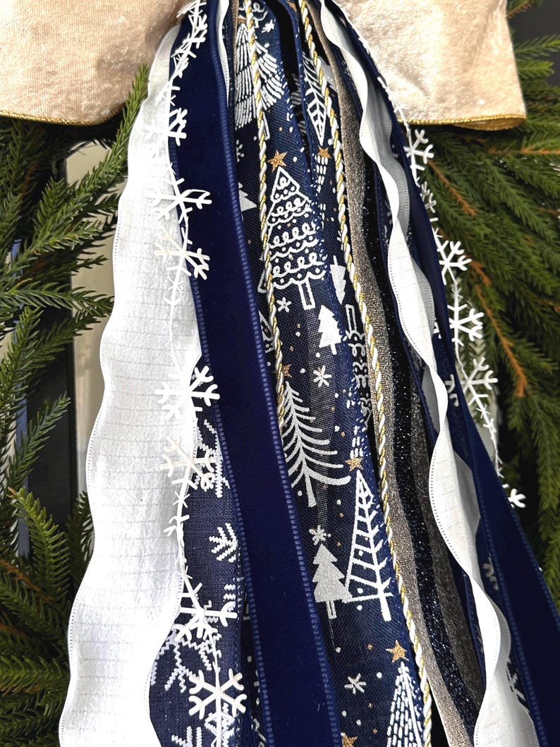 May include: A close-up of a Christmas wreath with various ribbons. The ribbons include white, navy blue, and silver, with snowflake and Christmas tree patterns. The wreath has a gold trim and is surrounded by green pine needles.