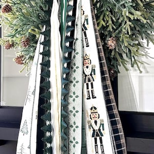 May include: A festive Christmas wreath with lush green pine branches, adorned with pinecones and decorative ribbons. The ribbons feature various patterns, including plaid, Christmas trees, and nutcracker soldiers, in shades of green, white, and gold.