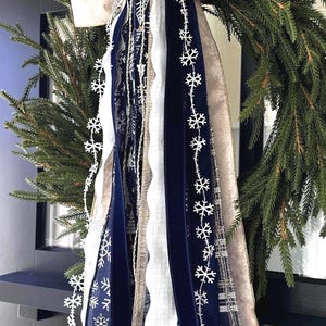 May include: A festive Christmas wreath with a large, cream-colored bow and cascading ribbons. The ribbons include navy blue velvet, white, and silver snowflake accents. The wreath is made of evergreen branches, perfect for holiday decor.