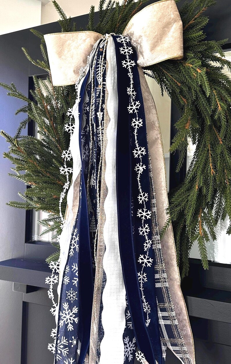 May include: A festive Christmas wreath with a large, cream-colored bow and cascading ribbons. The ribbons include navy blue velvet, white, and silver snowflake accents. The wreath is made of evergreen branches, perfect for holiday decor.