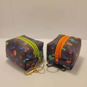 May include: Two small zippered pouches with a Halloween theme. The pouches are black with a colorful print of ghosts and jack-o-lanterns. Each pouch has a zipper with a contrasting color, one green and one orange. The pouches are attached to gold keychains.