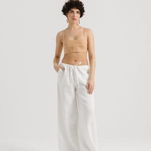 K&ouml;nnte beinhalten: Eine Frau tr&auml;gt eine wei&szlig;e Leinenhose mit Kordelzug und ein hellbraunes Crop-Top. Sie tr&auml;gt goldene Creolen und hat kurze, lockige braune Haare.