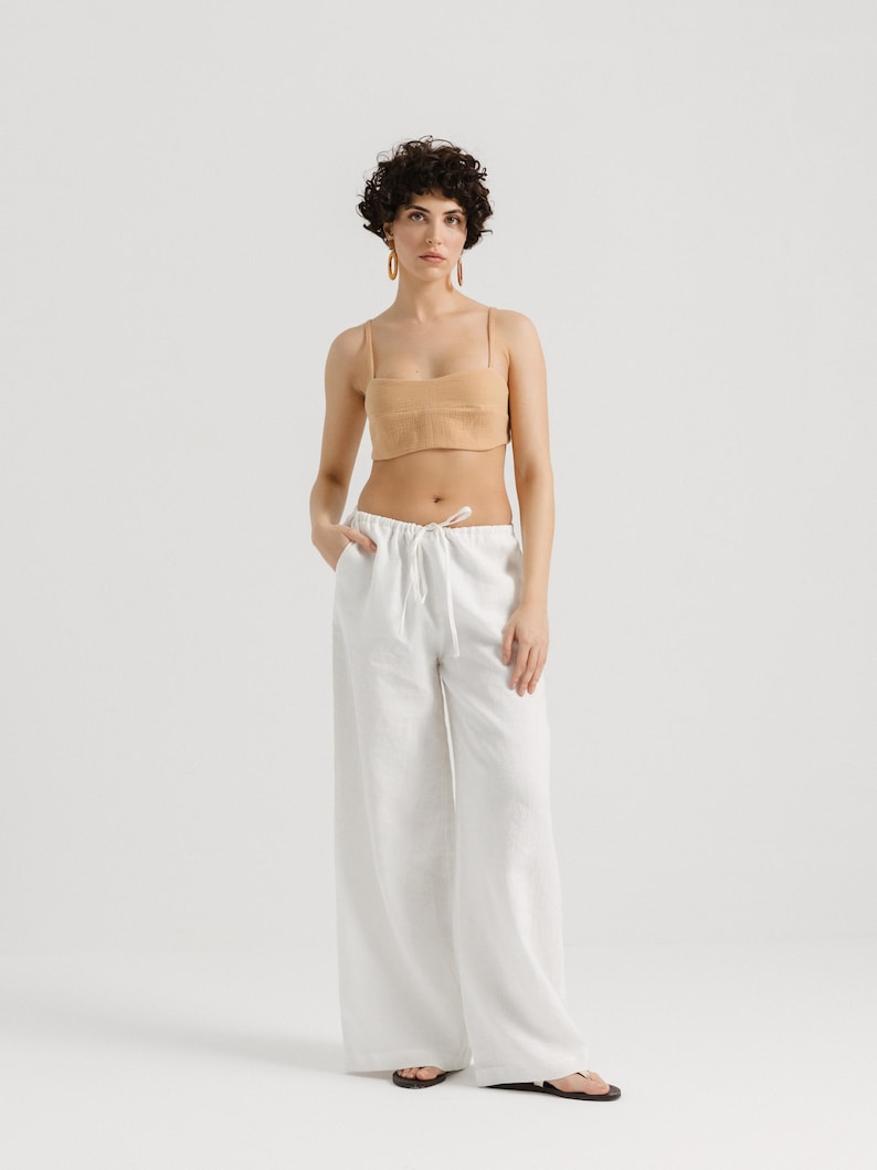 K&ouml;nnte beinhalten: Eine Frau tr&auml;gt eine wei&szlig;e Leinenhose mit Kordelzug und ein hellbraunes Crop-Top. Sie tr&auml;gt goldene Creolen und hat kurze, lockige braune Haare.